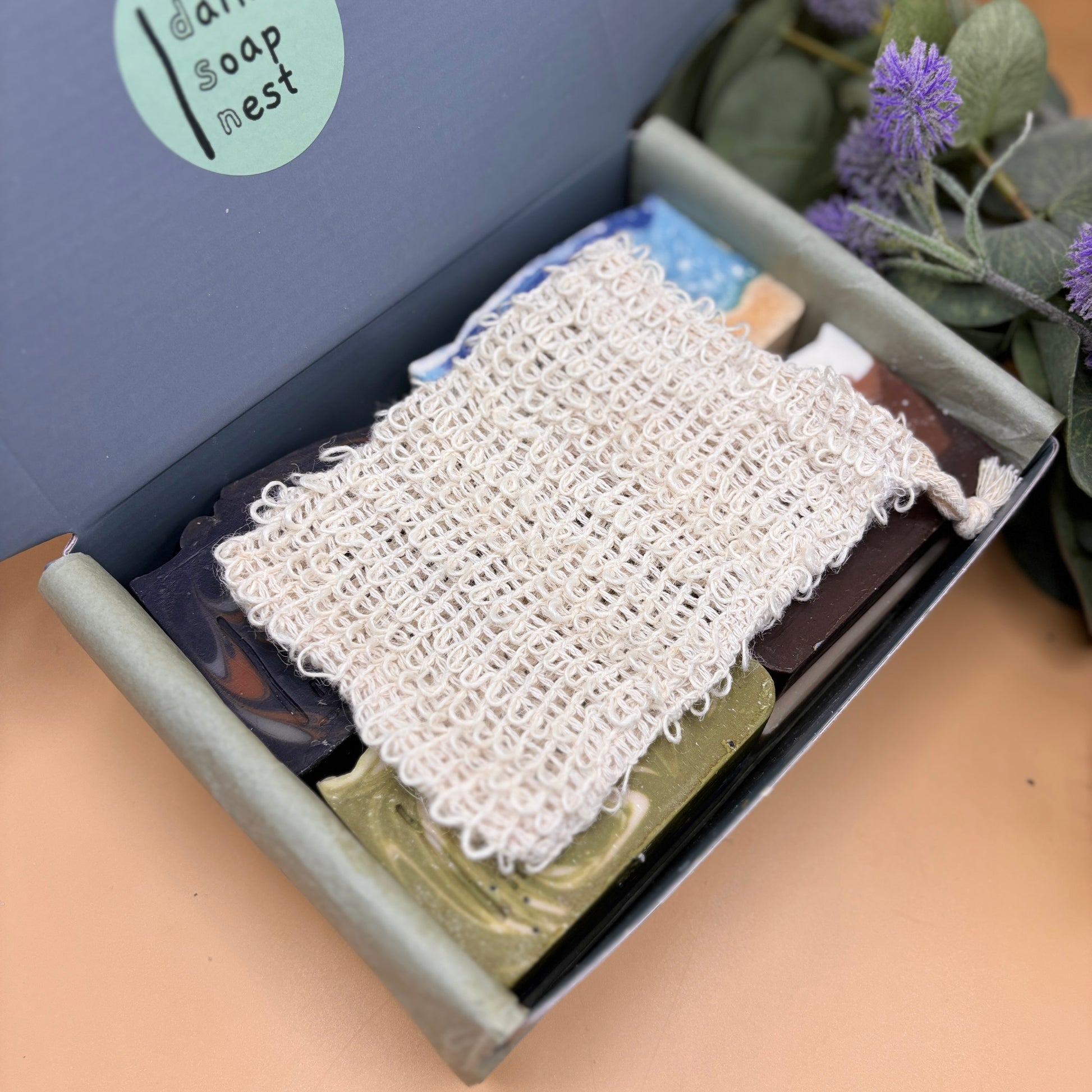 Box of soap with a white mesh scrubber and purple flowers on a beige surface